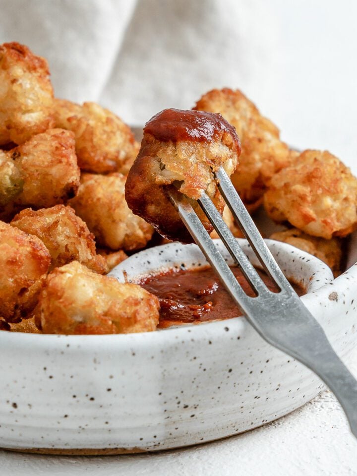 completed Air Fryer Tater Tots in a white speckled bowl with a fork in one being dipped in ketchup against a white background