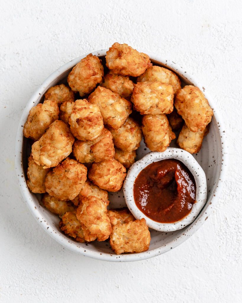 completed Air Fryer Tater Tots in a white speckled bowl and a small bowl of ketchup against a white background