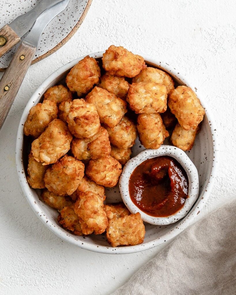 completed Air Fryer Tater Tots in a white speckled bowl and a small bowl of ketchup against a white background
