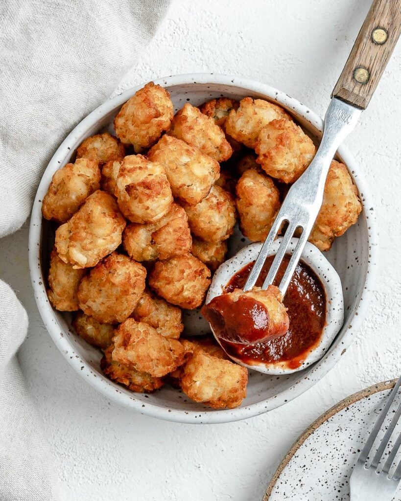 completed Air Fryer Tater Tots in a white speckled bowl with a fork in one being dipped in ketchup against a white background