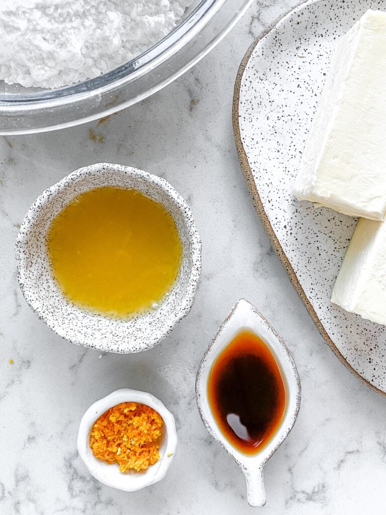 ingredients for Orange Buttercream Frosting measured out in individual white small bowls against a white marble background