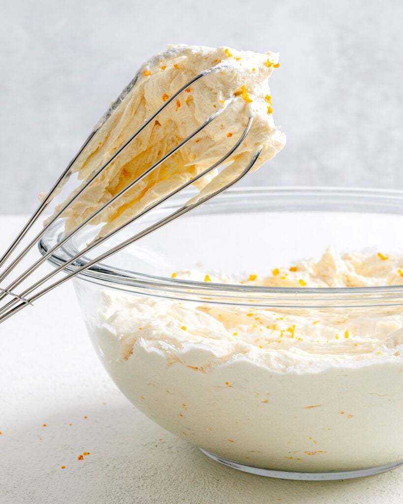completed Orange Buttercream Frosting in a glass bowl with a whisk against a white background