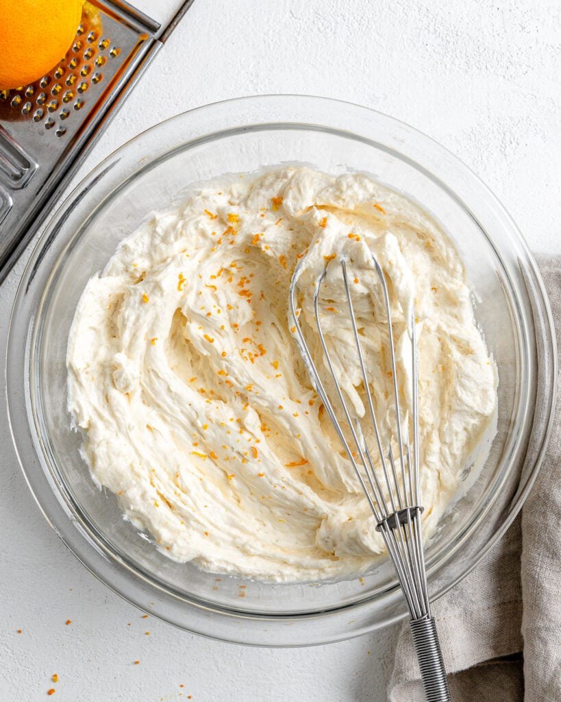 completed Orange Buttercream Frosting in a glass bowl with a whisk against a white background with an orange in the background