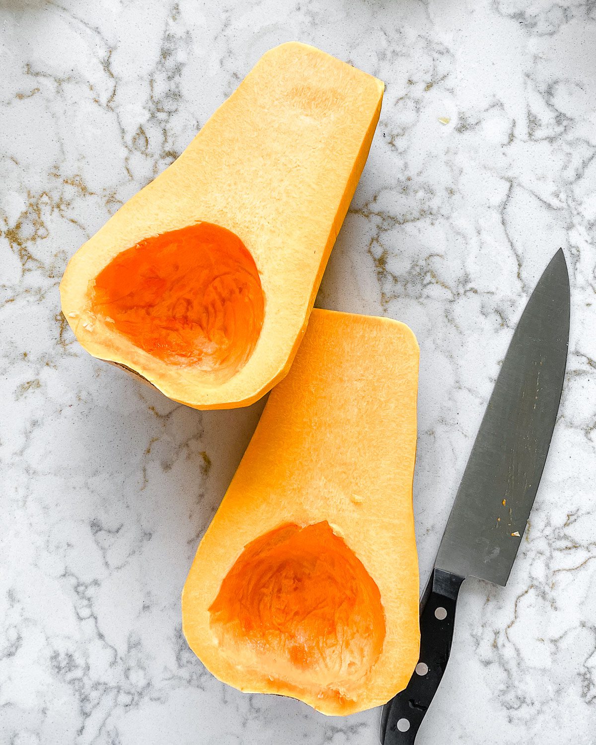 halved butternut squash with a knife laid out against a white marble background