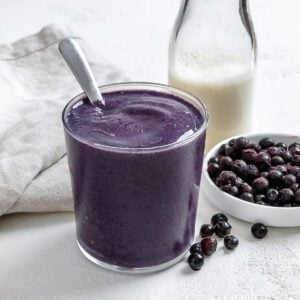 completed Blueberry Banana Smoothie in a glass with blueberries and plant based milk in the background