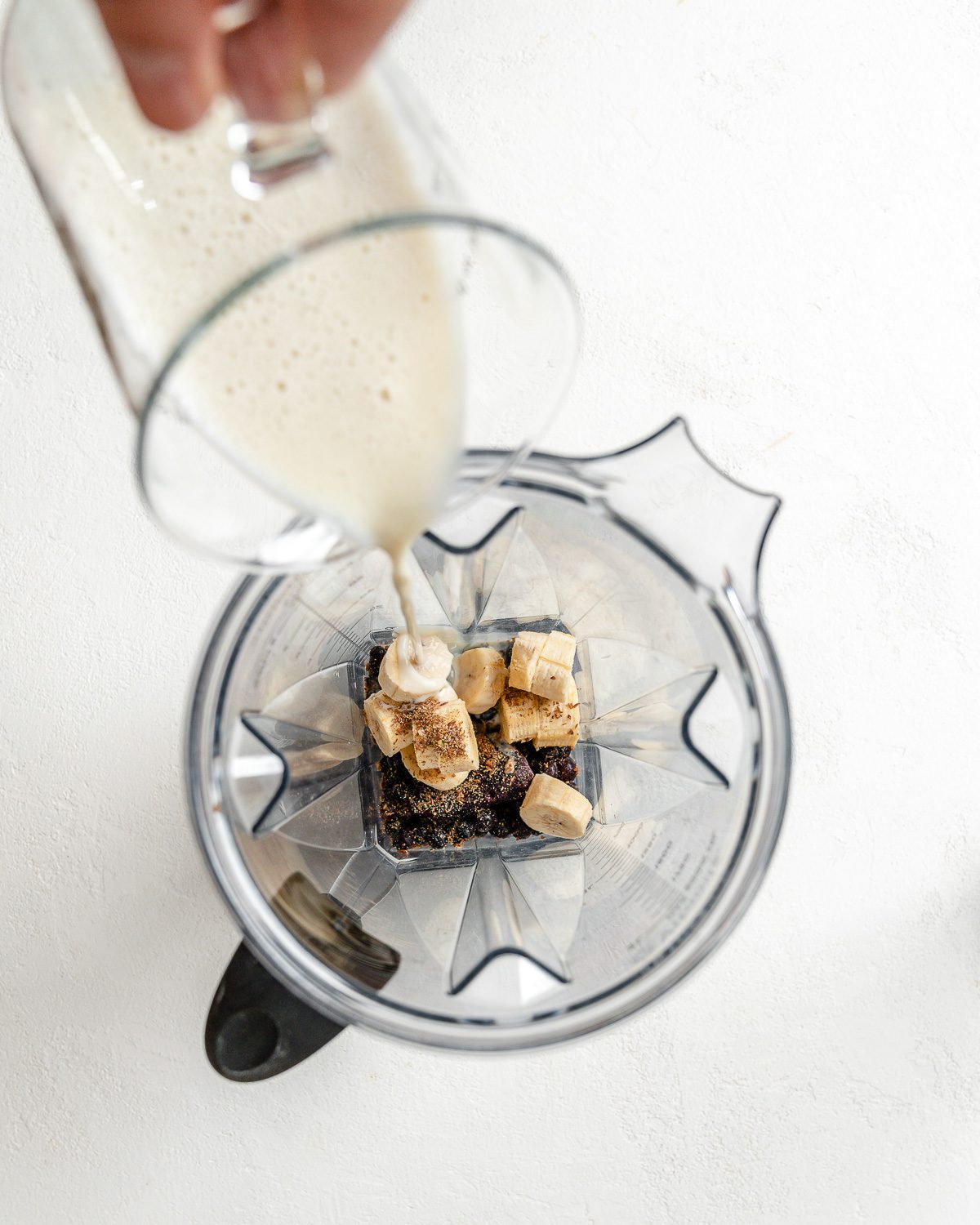 process of pouring plant milk into blender with fruits against a white background