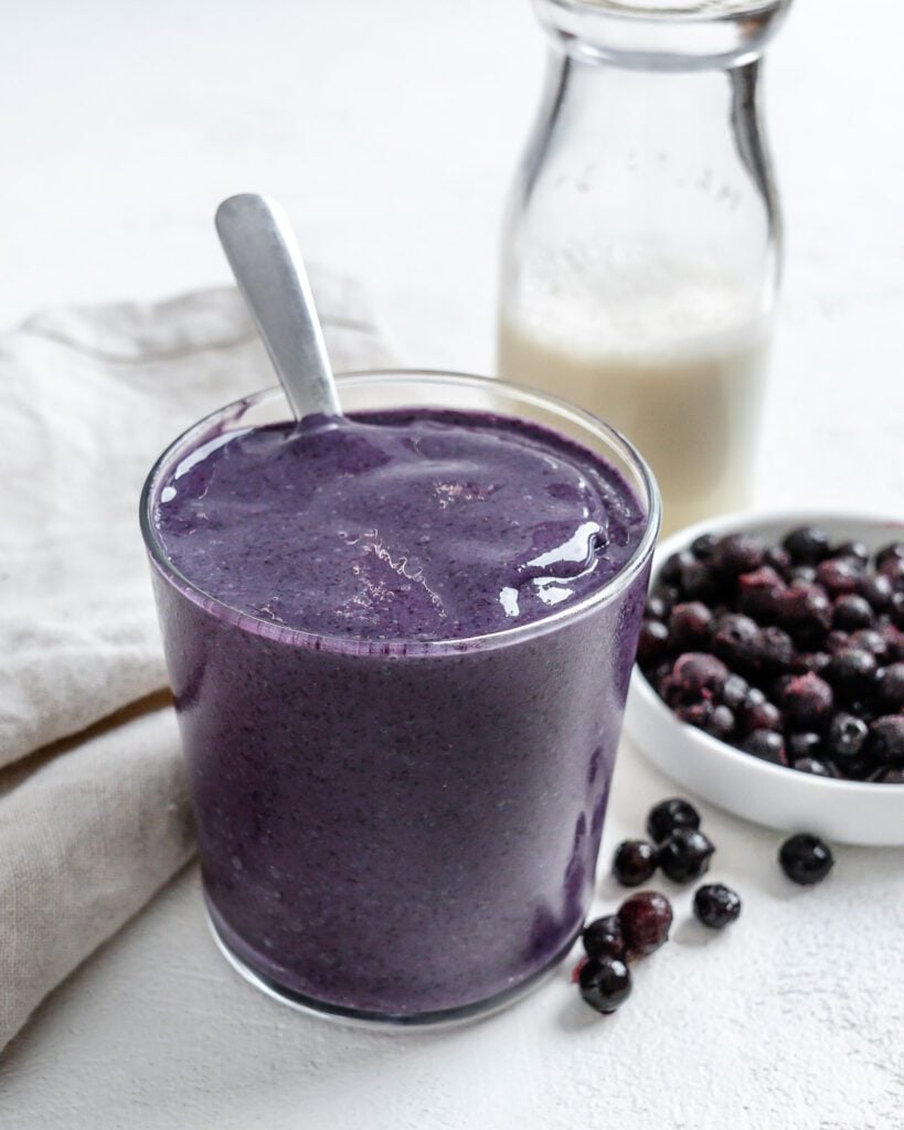 completed Blueberry Banana Smoothie in a glass with blueberries and plant based milk in the background