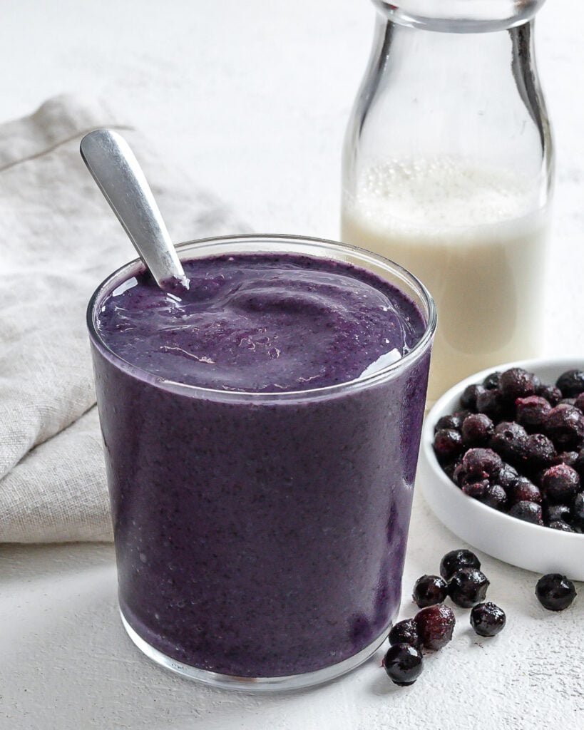 completed Blueberry Banana Smoothie in a glass with blueberries and plant based milk in the background