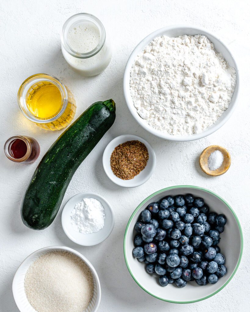 ingredients for blueberry zucchini muffins measured out in individual bowls against a white backgorund