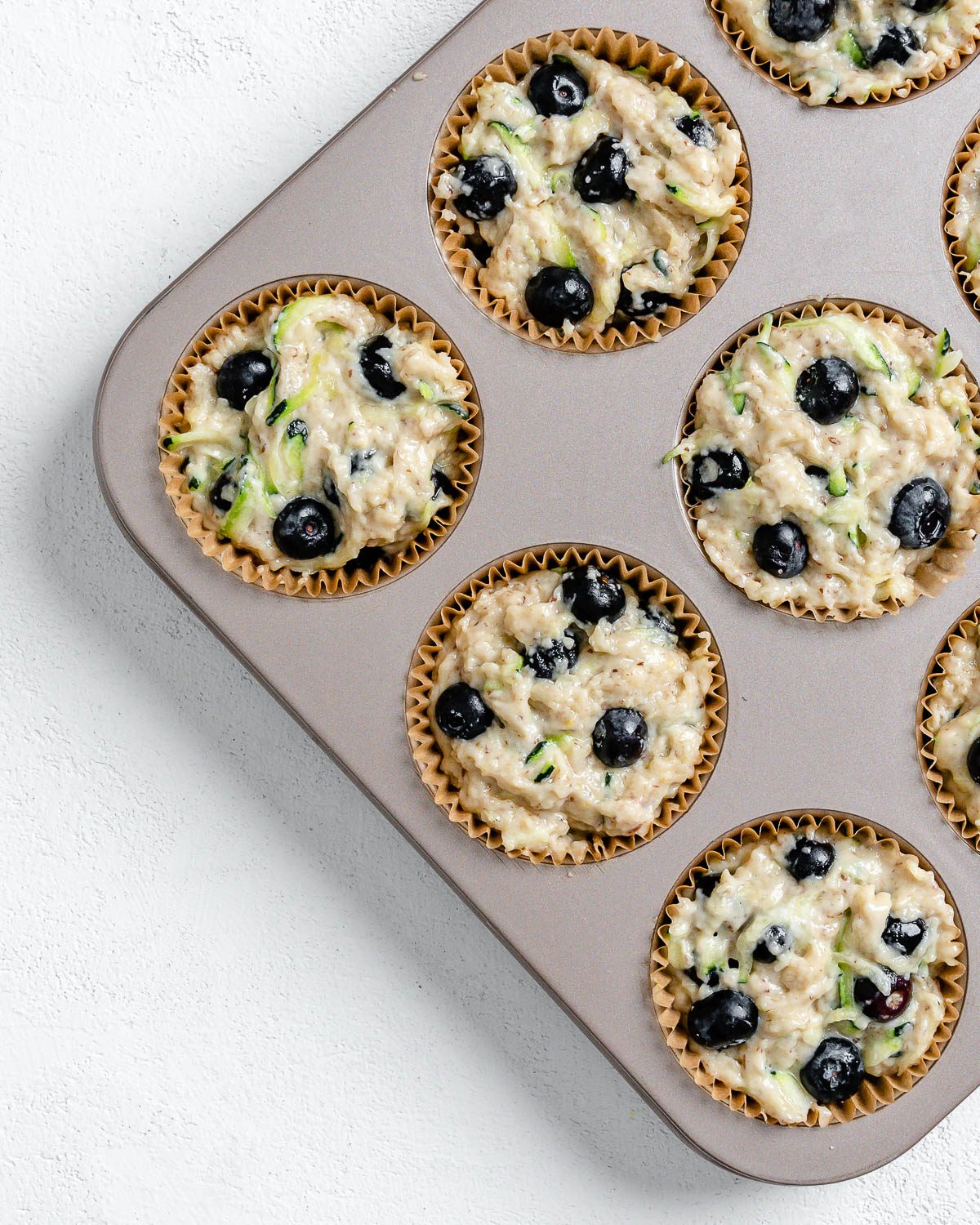 process of adding batter and blueberries to the top of batter in a muffin tin against a white background