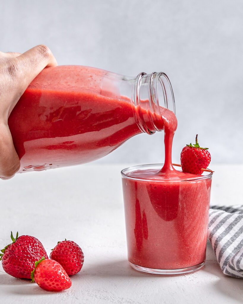 process of pouring completed strawberry juice from a jar into a cup with scattered strawberries against a white background