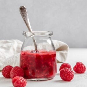 completed Vegan Raspberry Syrup in a glass jar with raspberries and a white cloth against a white background