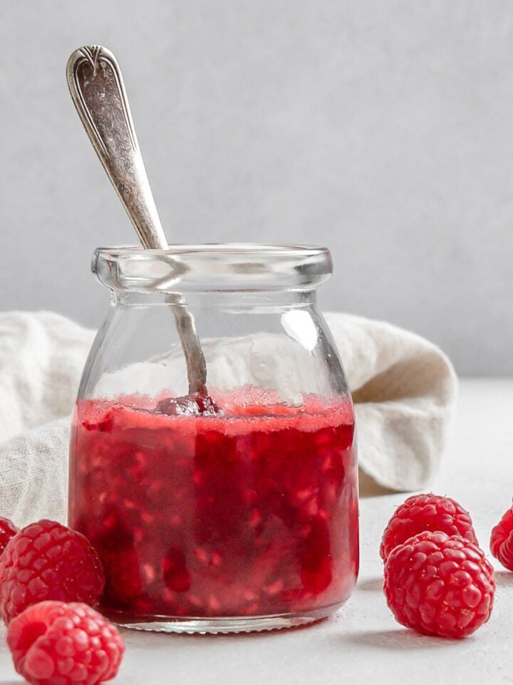 completed Vegan Raspberry Syrup in a glass jar with raspberries and a white cloth against a white background