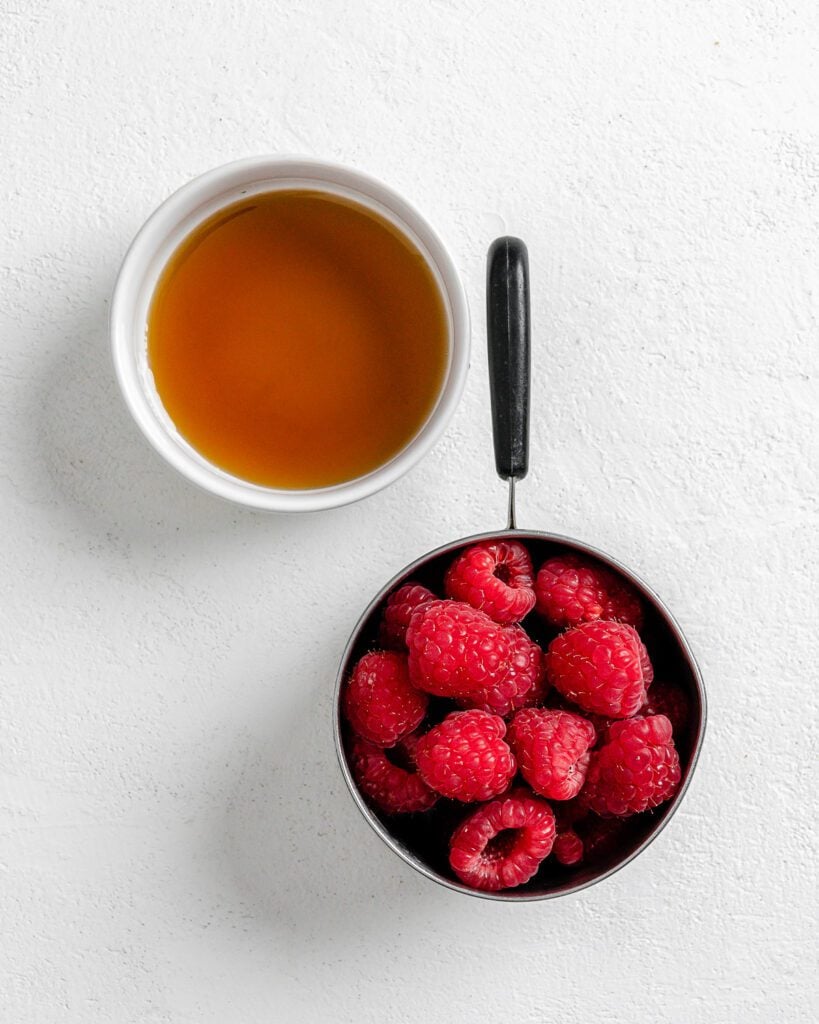 ingredients of raspberries in a measuring cup and liquid ingredient in white bowl against white background