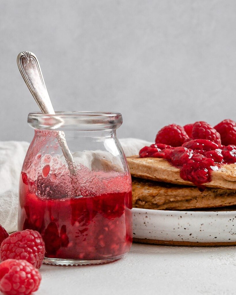 completed Vegan Raspberry Syrup in a glass jar and on top of a stack of pancakes against a white surface