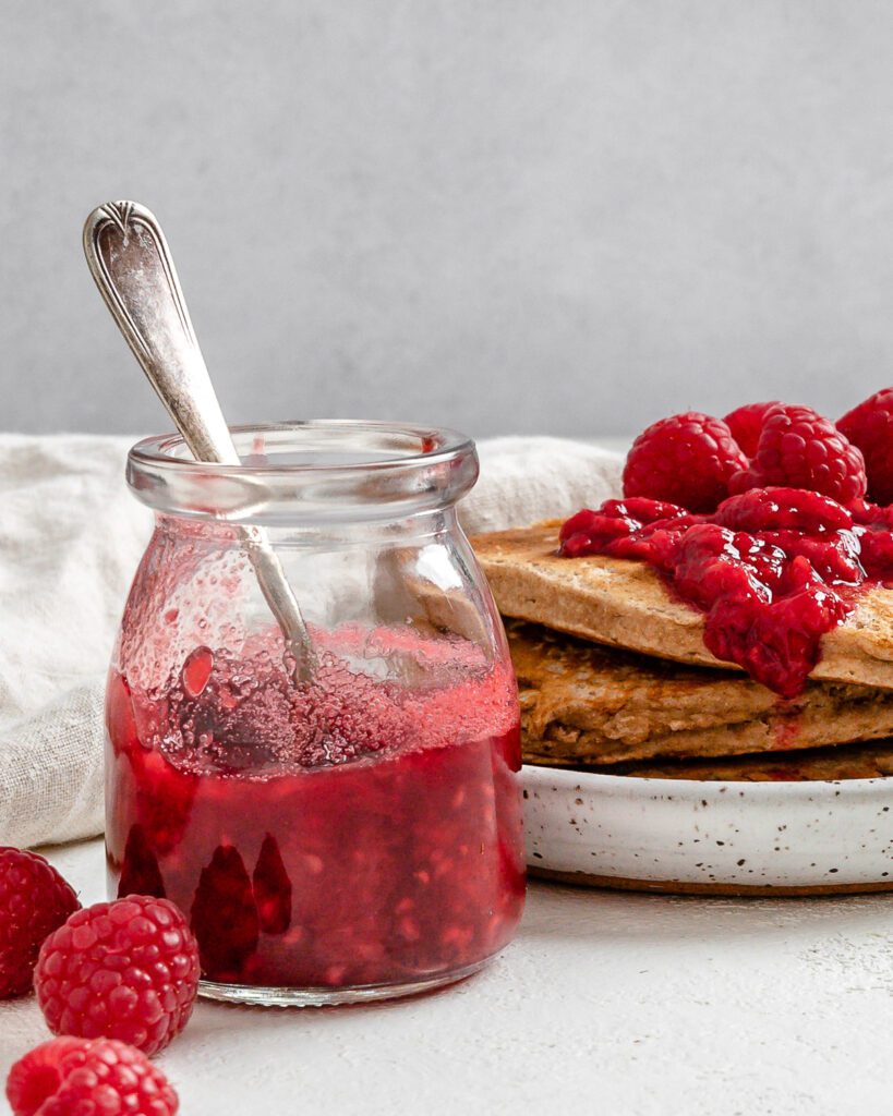 completed Vegan Raspberry Syrup in a glass jar and on top of a stack of pancakes against a white surface