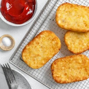 four completed hash brown patties on a air fryer tray with ketchup and salt in the background