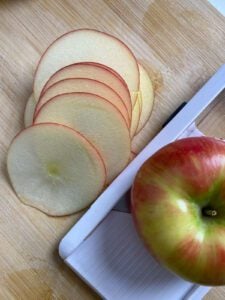 Thin round apple slices on a wooden cutting board next to a mandoline and a whole apple.