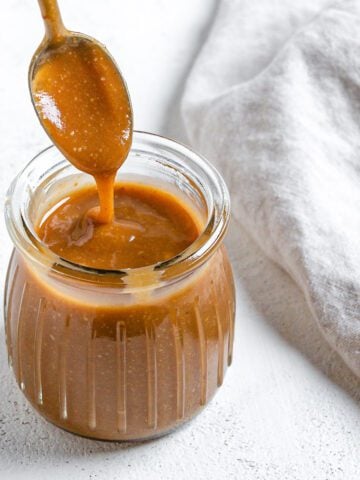 completed Simple Vegan Caramel in a jar with caramel drizzling down from a spoon against a white background
