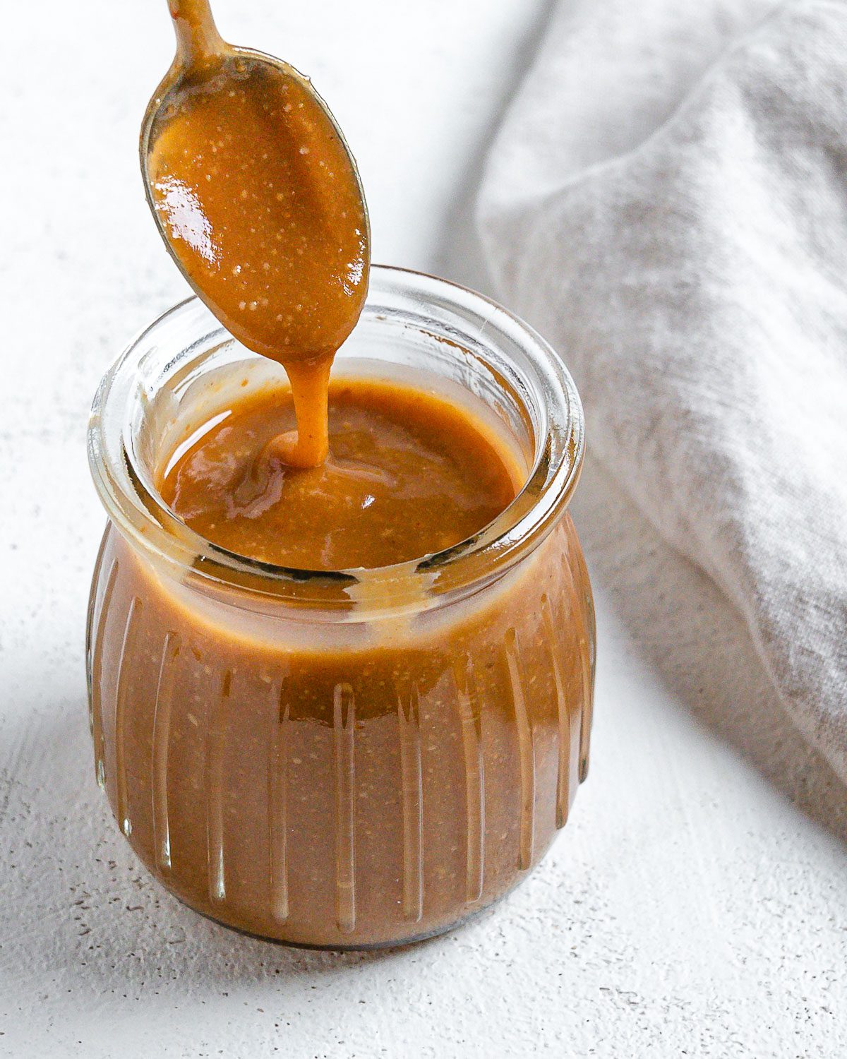 completed Simple Vegan Caramel in a jar with caramel drizzling down from a spoon against a white background