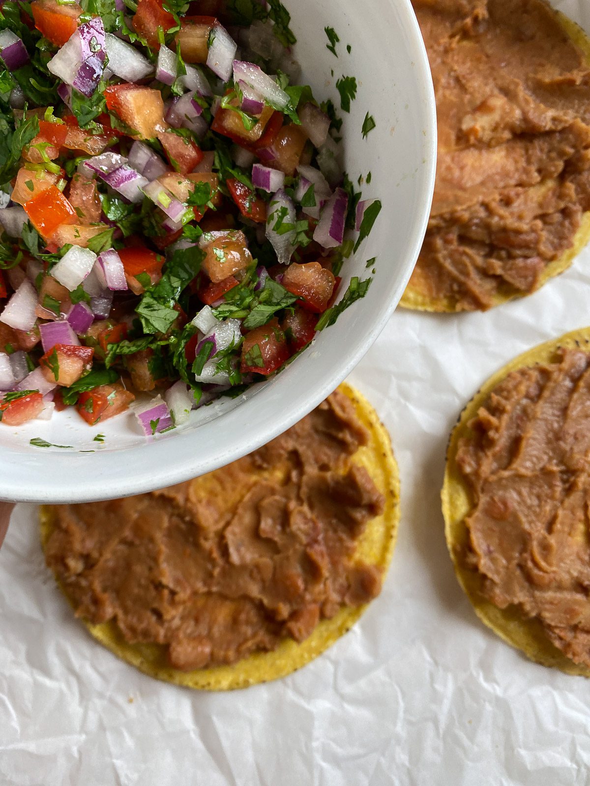 process of veggies being added on top of refried beans onto tostada shell