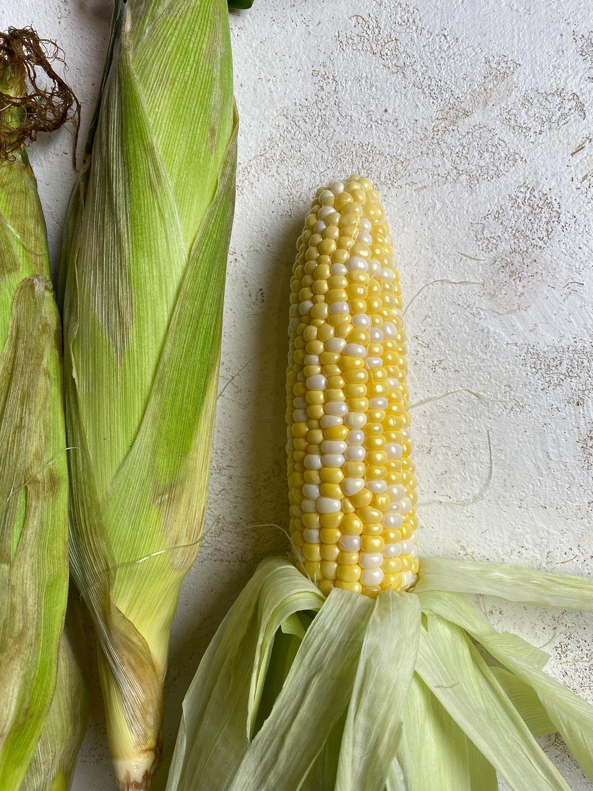three cobs of corn with one peeled on a light surface