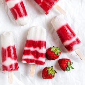 several completed Vegan Strawberry Popsicles on a white surface with strawberries alongside of them