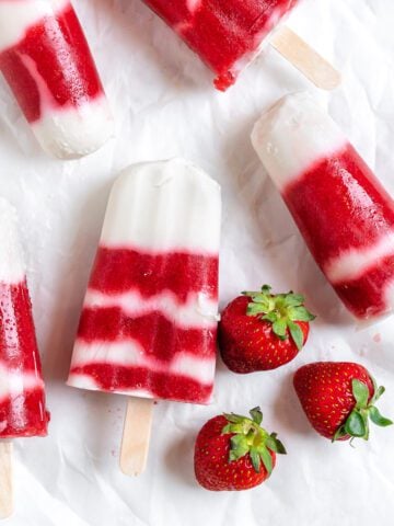 several completed Vegan Strawberry Popsicles on a white surface with strawberries alongside of them