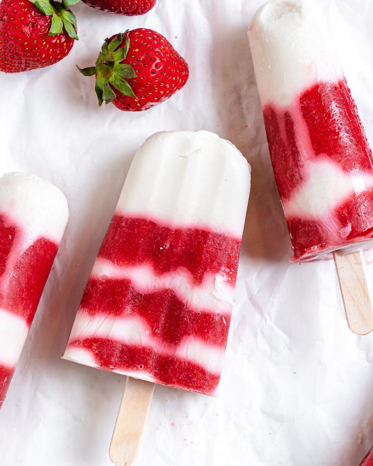 several completed Vegan Strawberry Popsicles on a white surface with strawberries alongside of them