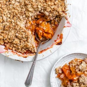 completed Vegan Peach Crisp in a white baking dish and with a piece plated on a plate against a white background