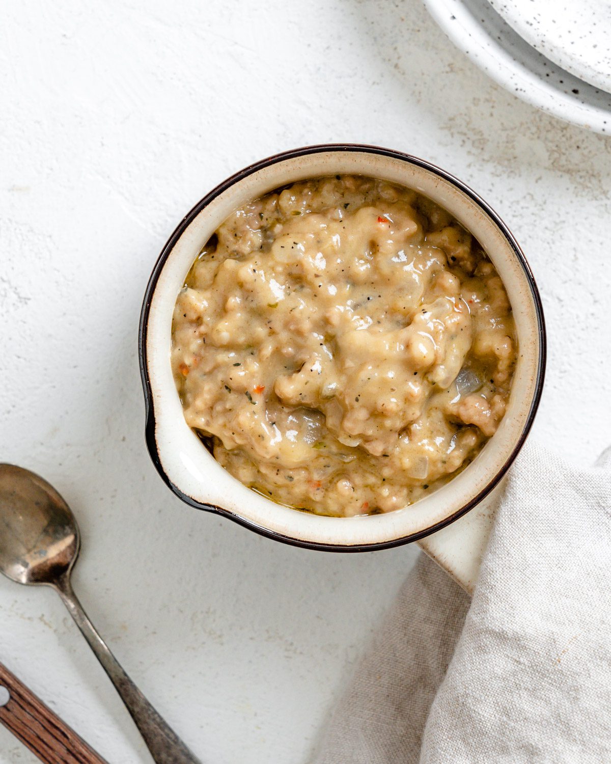 Overhead view of vegan sausage gravy in a small cream bowl with a spout, a spoon and a linen napkin beside it.