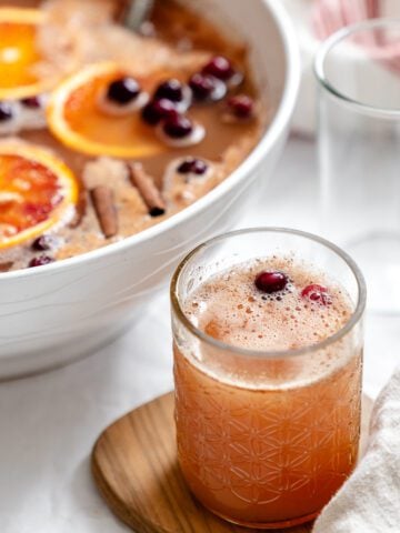 A glass of non-alcoholic Thanksgiving punch with a cranberry on top, in front of the punch bowl.