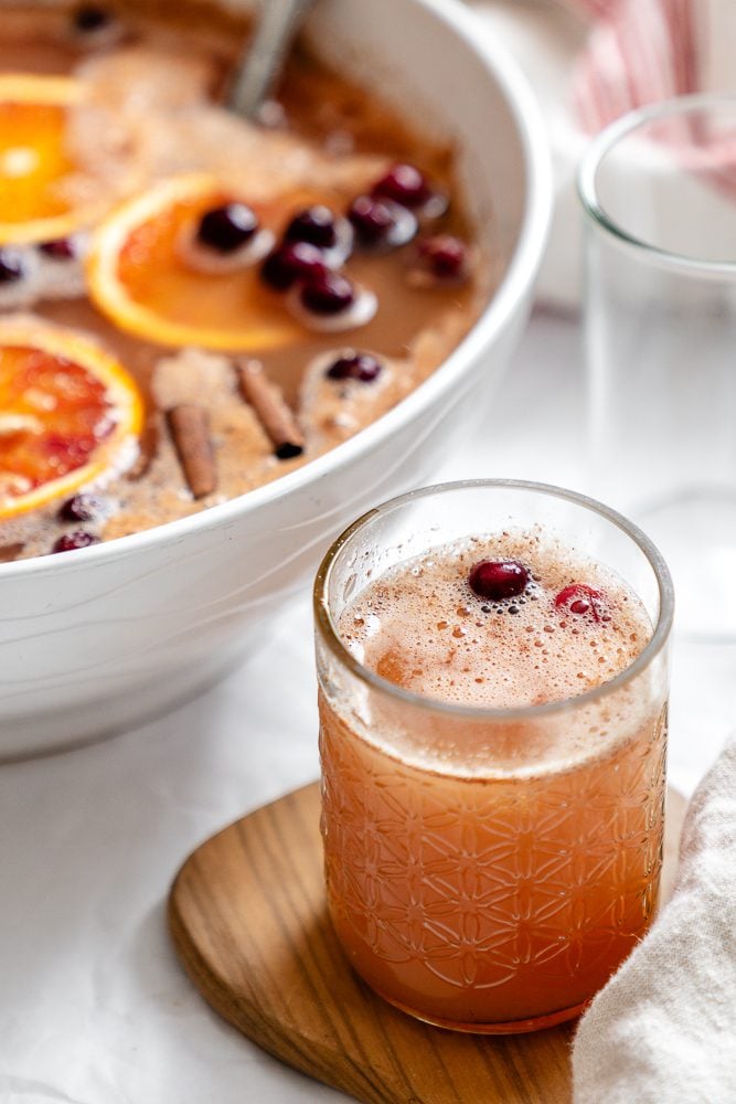 A glass of non-alcoholic Thanksgiving punch with a cranberry on top, in front of the punch bowl.