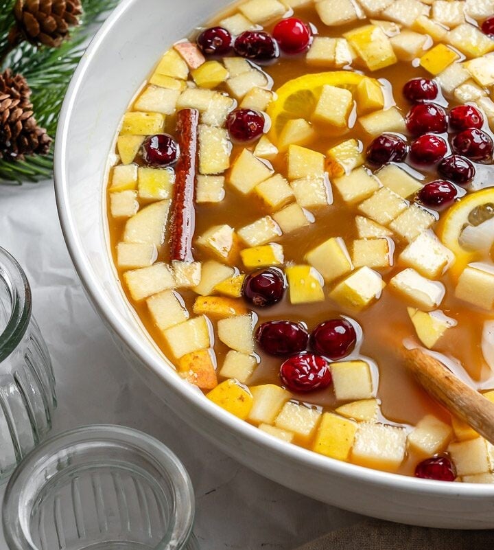 Bowl of non-alcoholic Christmas punch with diced apple, cranberries, lemon slices, and a cinnamon stick, next to empty glasses, pine branches, and ornaments.